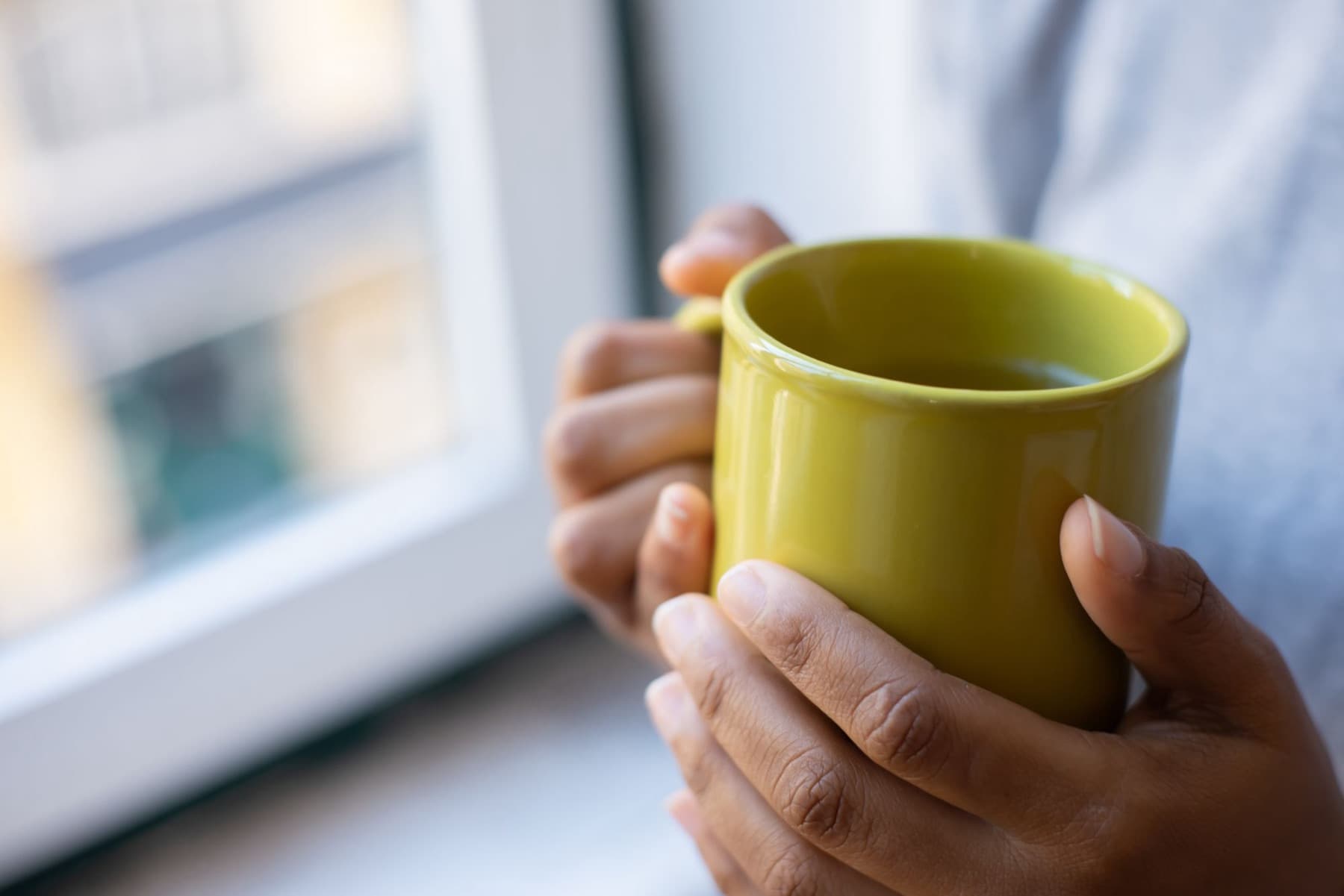 Two hands hold a yellow green mug of tea beside a bright window