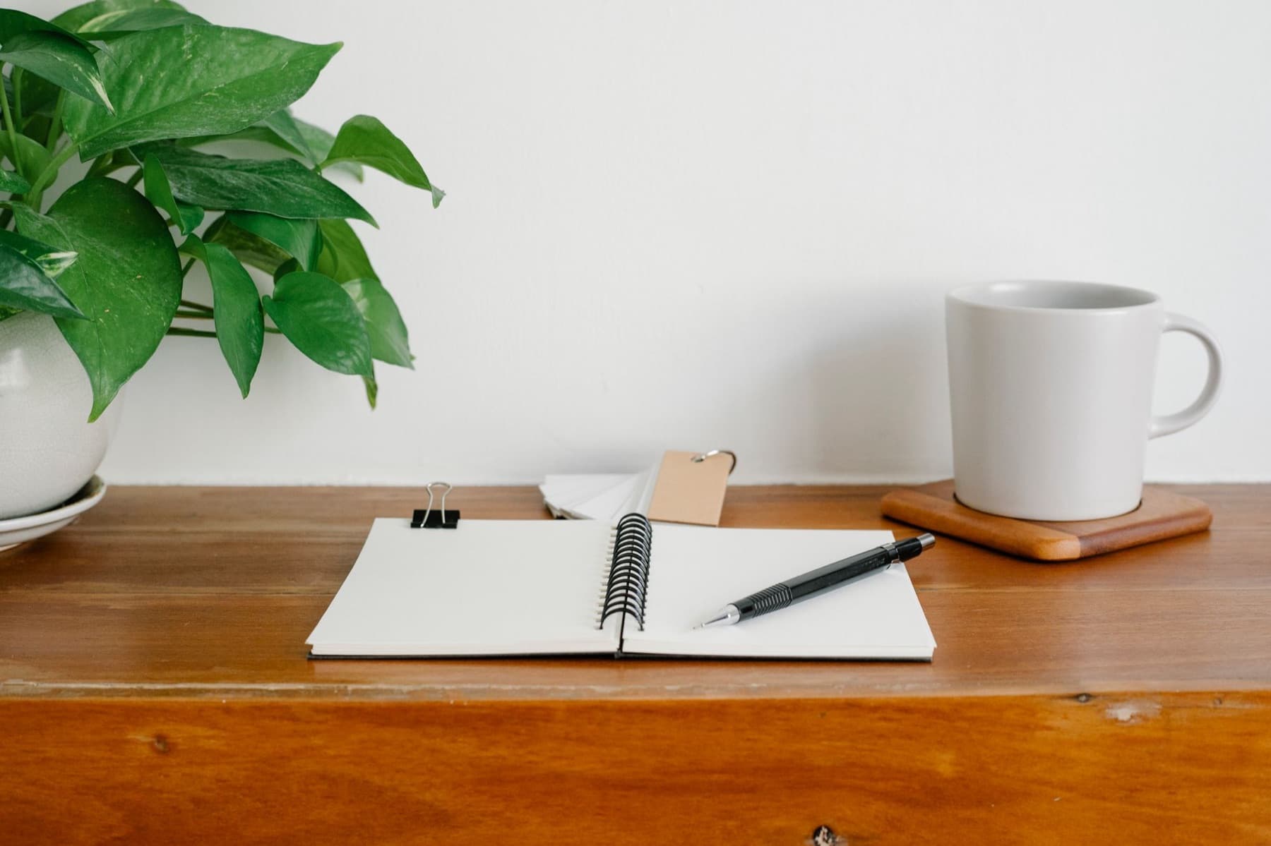 An open notebook with a pencil, a white mug on a wooden coaster and a pot plant on a wooden desk