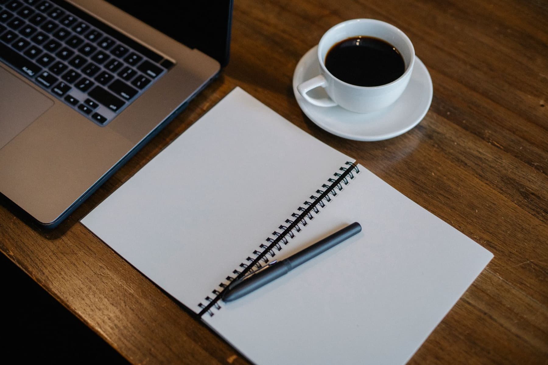 A laptop, an open spiral notebook with a pen and a cup of black coffee on a wooden desk