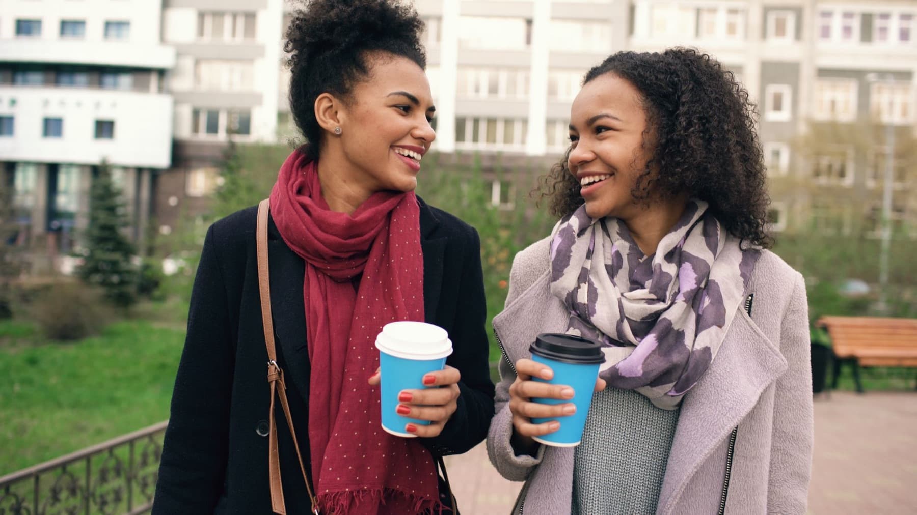 Two women laugh together while walking outdoors with takeaway coffee cups