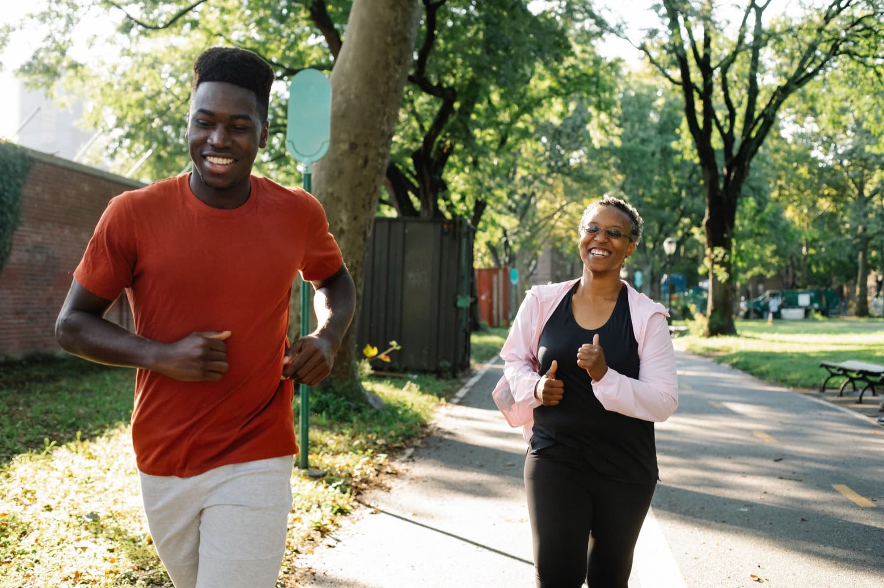 A man and a woman jog side by side along a tree lined park path on a sunny day