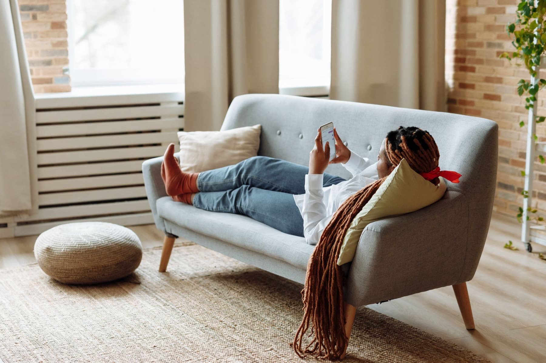 A woman lies back on a grey sofa in a bright living room, looking at her phone