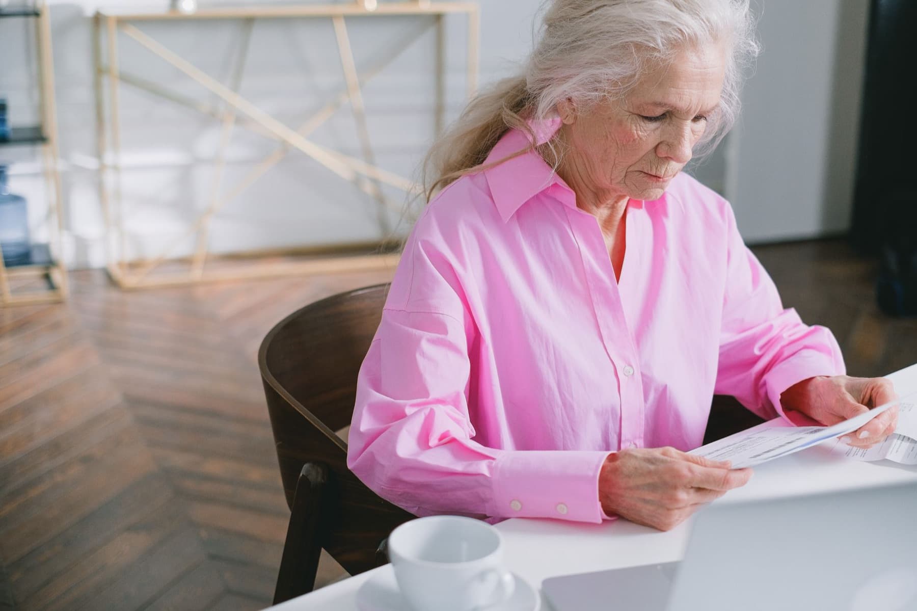 An older woman in a pink shirt reads a printed letter at a table next to a cup and a laptop